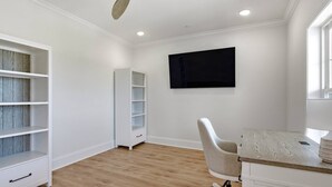 A room with a ceiling fan, a TV mounted on the wall, a desk with a chair, and built-in shelving. - Brand-New Oceanfront Showstopper | 6 En-Suite Bedrooms, Pool, 2 Hot Tubs, Elevator (North Myrtle Beach)