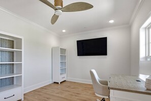 Ein Zimmer mit Deckenventilator, einem an der Wand montierten Fernseher, Schreibtisch mit Stuhl und fest installierten Regalen.