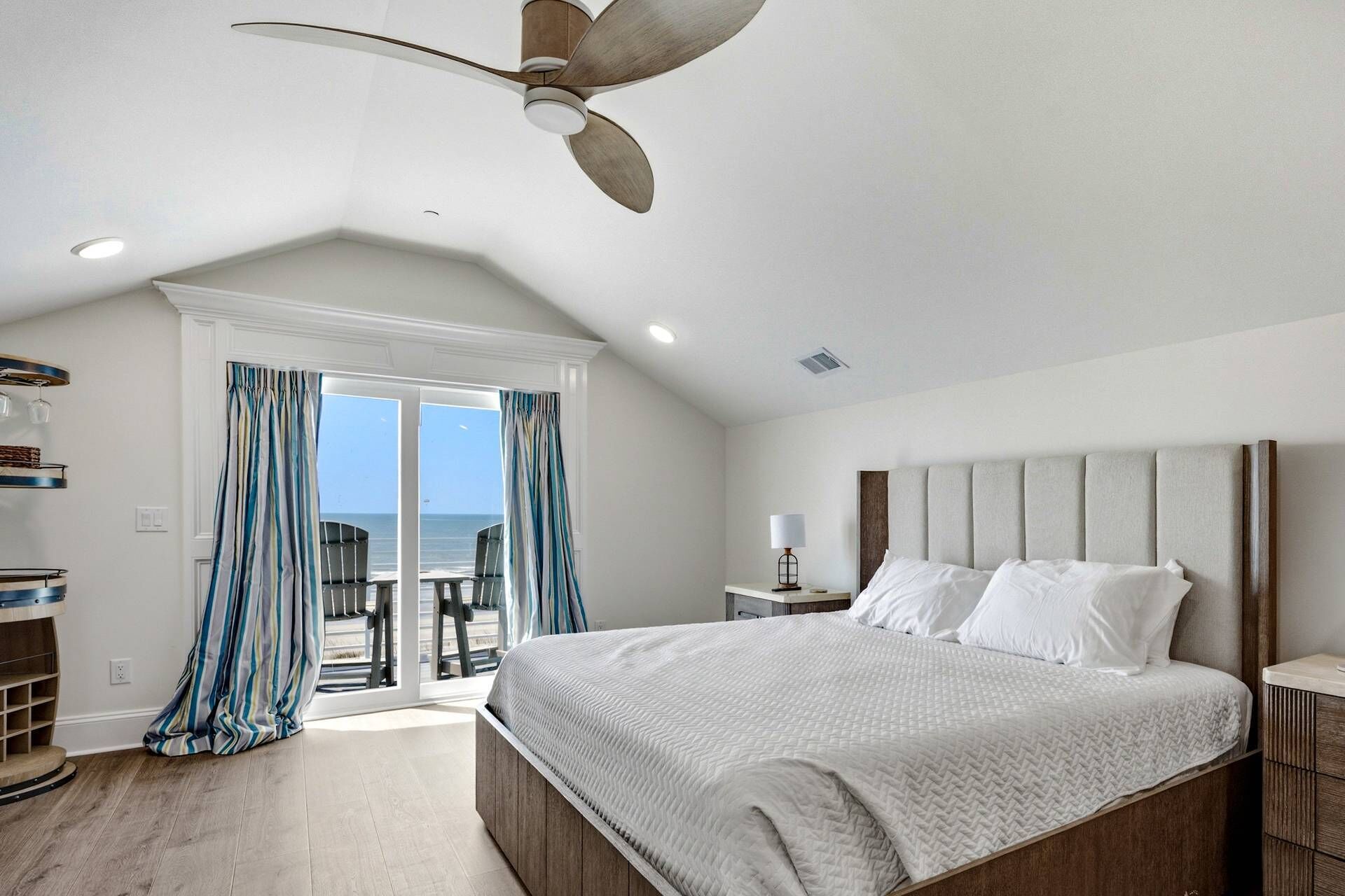 A bedroom with a large bed, a ceiling fan, a nightstand, and a view of the ocean through a sliding door.