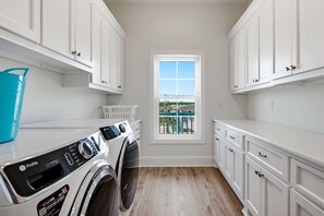 A laundry room with a front-loading washer and dryer, white cabinets, a window with a view, and a blue laundry basket. - Brand-New Oceanfront Showstopper | 6 En-Suite Bedrooms, Pool, 2 Hot Tubs, Elevator (North Myrtle Beach)