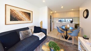 A modern living room with a dark sofa, a dining area with a round table and chairs, and a kitchen with light wooden cabinets.