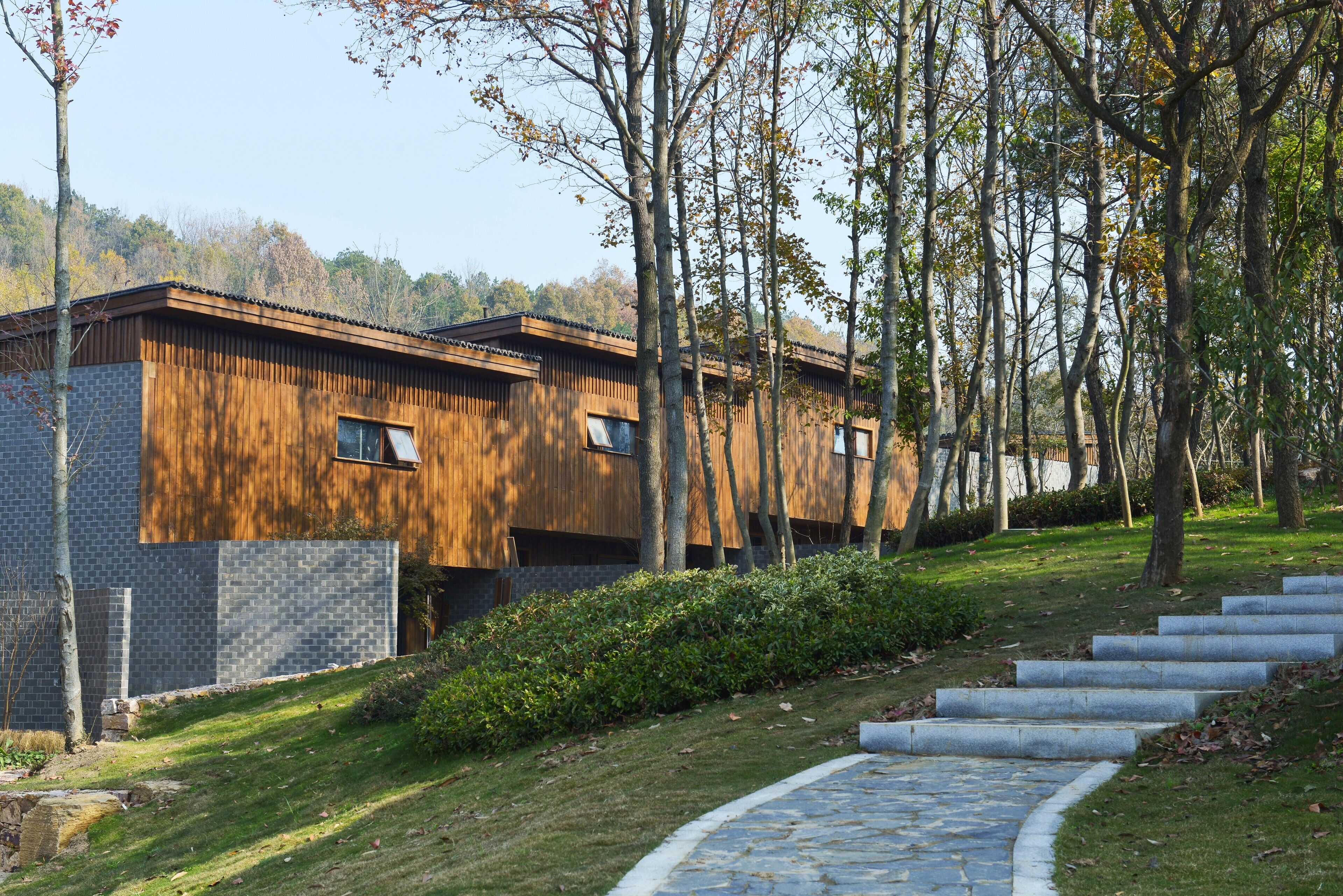 A modern building with a wooden facade and stone base, surrounded by trees and a stone pathway.
