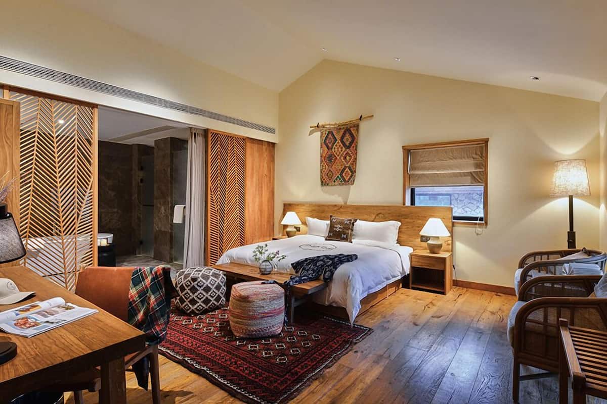 Un dormitorio con cama, mesitas de noche, suelo de madera, ventana y vista a un muro de piedra.
