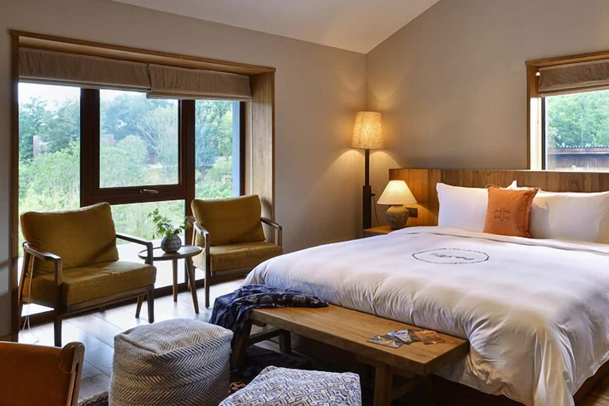 Habitación de hotel con una cama grande, dos lámparas de noche, un cabecero de madera, una mesita, una silla y vistas a zonas verdes a través de las ventanas.