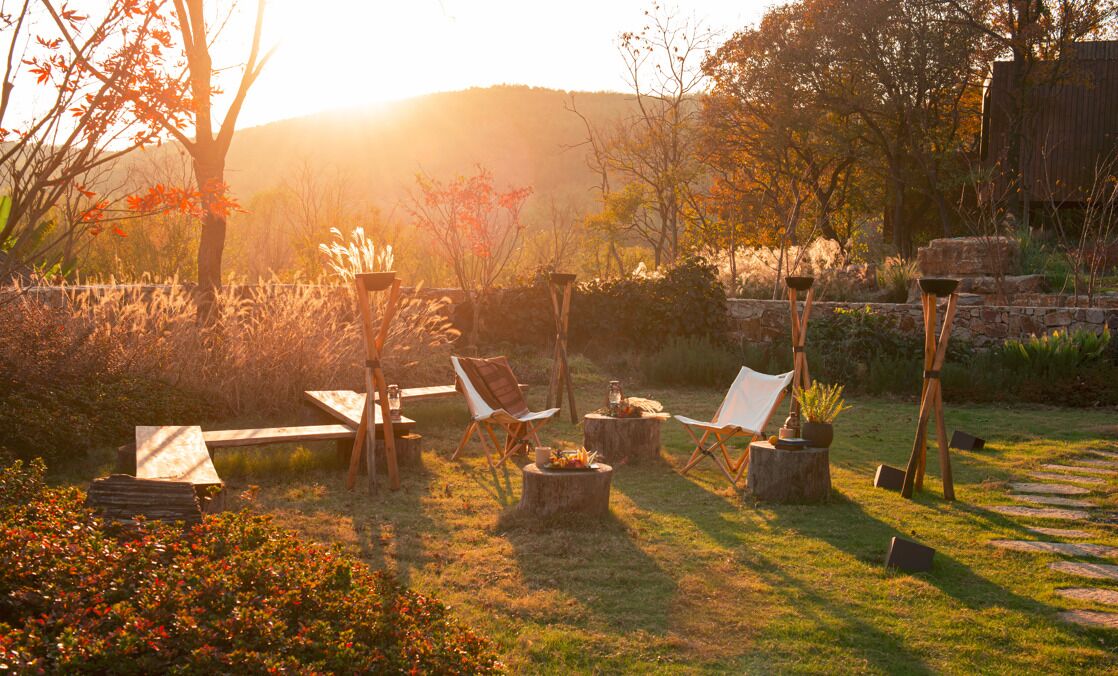 An outdoor seating area with wooden chairs and tables, surrounded by greenery and a stone wall.