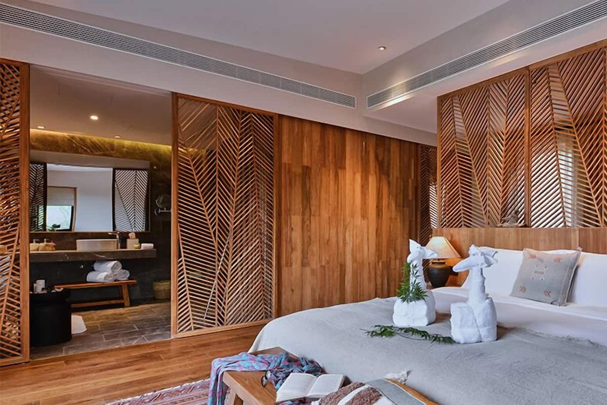 Una habitación de hotel moderna con una cama grande, paneles de madera, un baño con espejo y un banco.