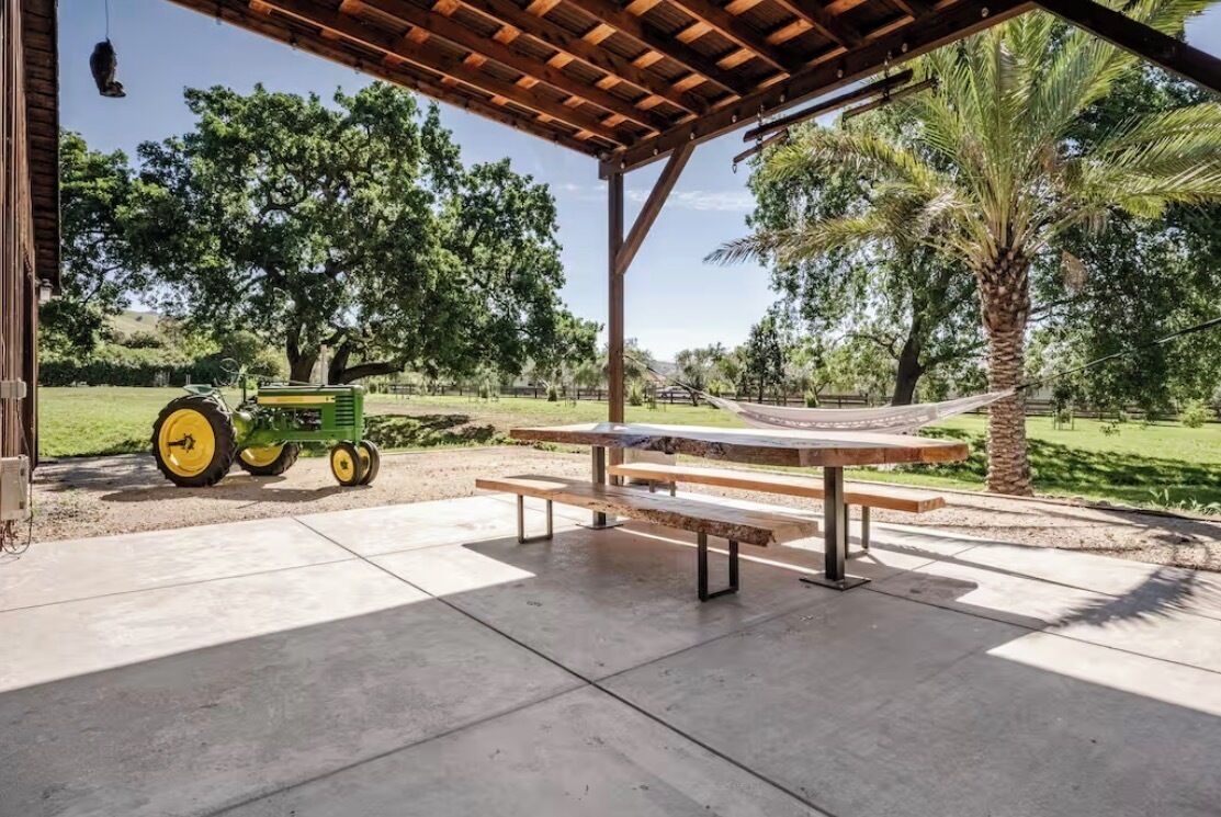 Un espacio al aire libre techado con una mesa de picnic y bancos, una hamaca y un tractor verde.
