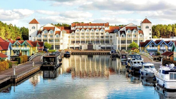 Un hotel frente a un canal, con un edificio central, barcos amarrados a lo largo del agua y casas coloridas a ambos lados.