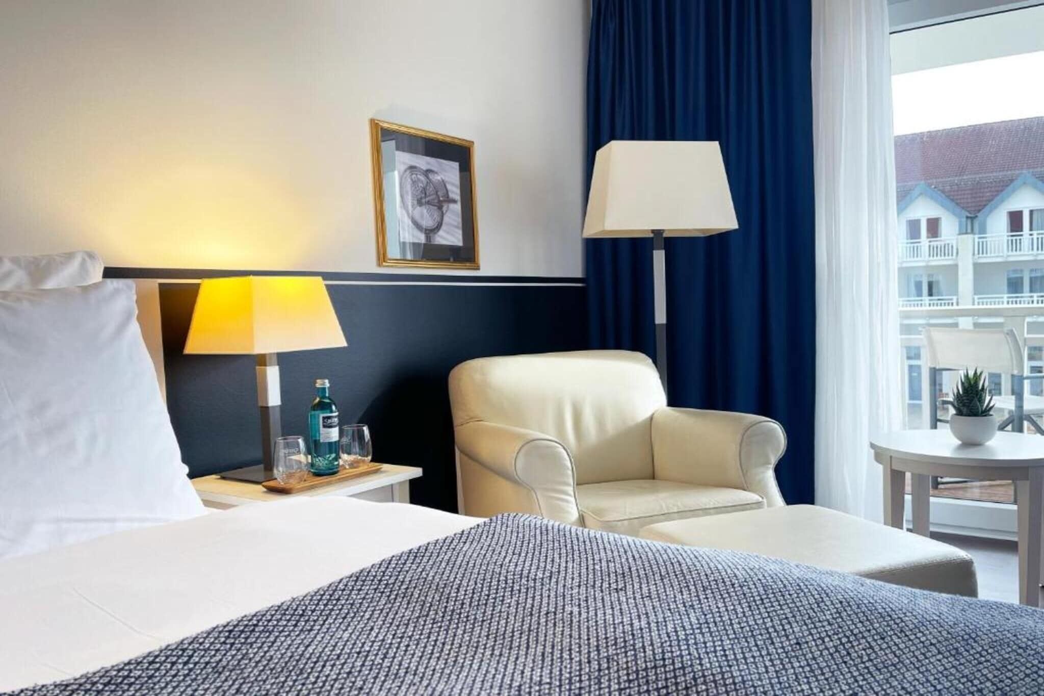 A hotel room with a bed, a chair, a nightstand with a lamp, a bottle of water, and a framed picture on the wall.