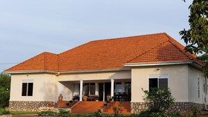 A two-story house with a stone foundation, a red-tiled roof, and a covered porch with outdoor furniture. - Martha's Barn (Mukono)