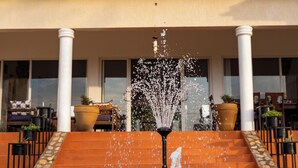 A fountain with water jets in front of a building with columns and potted plants. - Martha's Barn (Mukono)