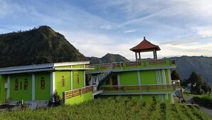 A green building with a red-roofed pavilion, surrounded by lush greenery and mountains. - Surya Bromo Homestay (Ngadisari)