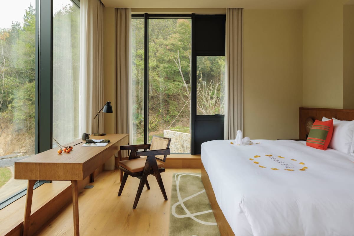 Habitación de hotel con una cama grande, un escritorio y una silla. Se ve una ventana con vista a árboles.
