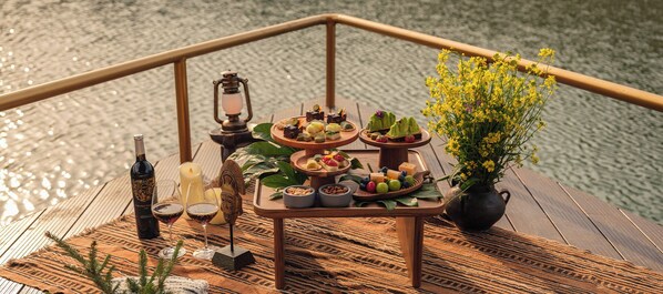 A wooden deck with a table set for a meal, including fruits, cheeses, and a bottle of wine.
