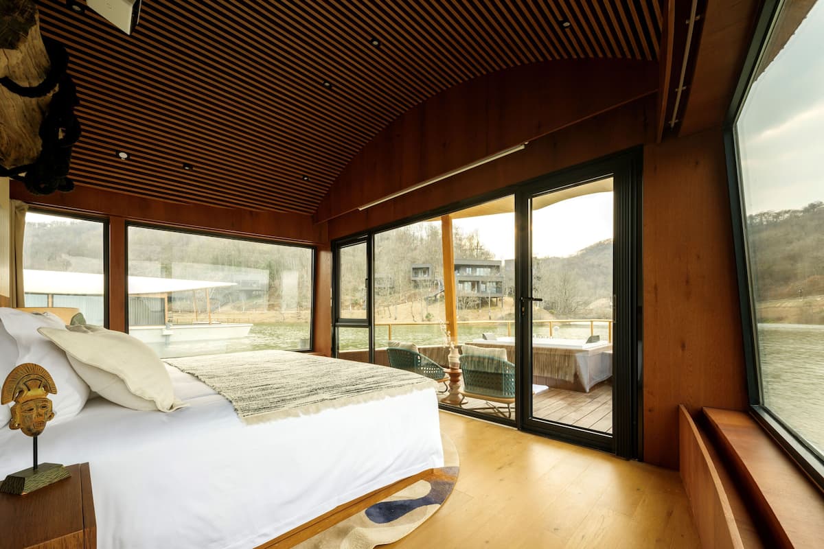 Un dormitorio con una cama grande, techo de madera y vistas a un lago y edificios a través de amplios ventanales.