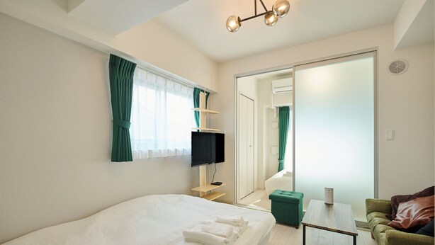 A hotel room with a large bed, a TV, a small table, a green chair, and a sliding door leading to another room. - Gekkō Hakata Higashi (Fukuoka)