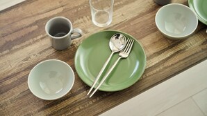 A wooden table with a green plate, a spoon, a fork, and two gray cups. - Gekkō Hakata Higashi (Fukuoka)
