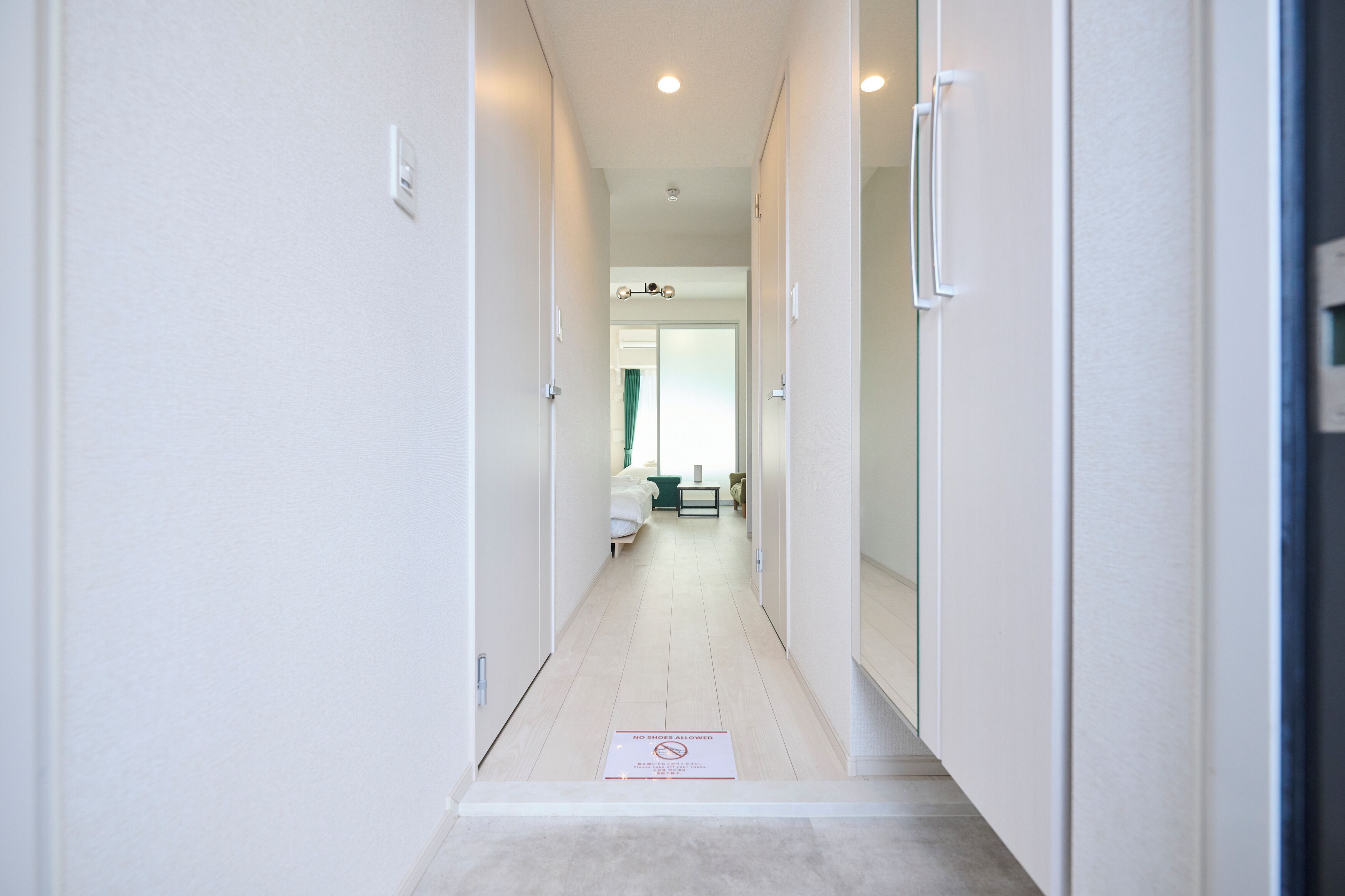 A narrow hallway with a 'No Shoes Allowed' sign, leading to a bathroom with a shower curtain and a bed.