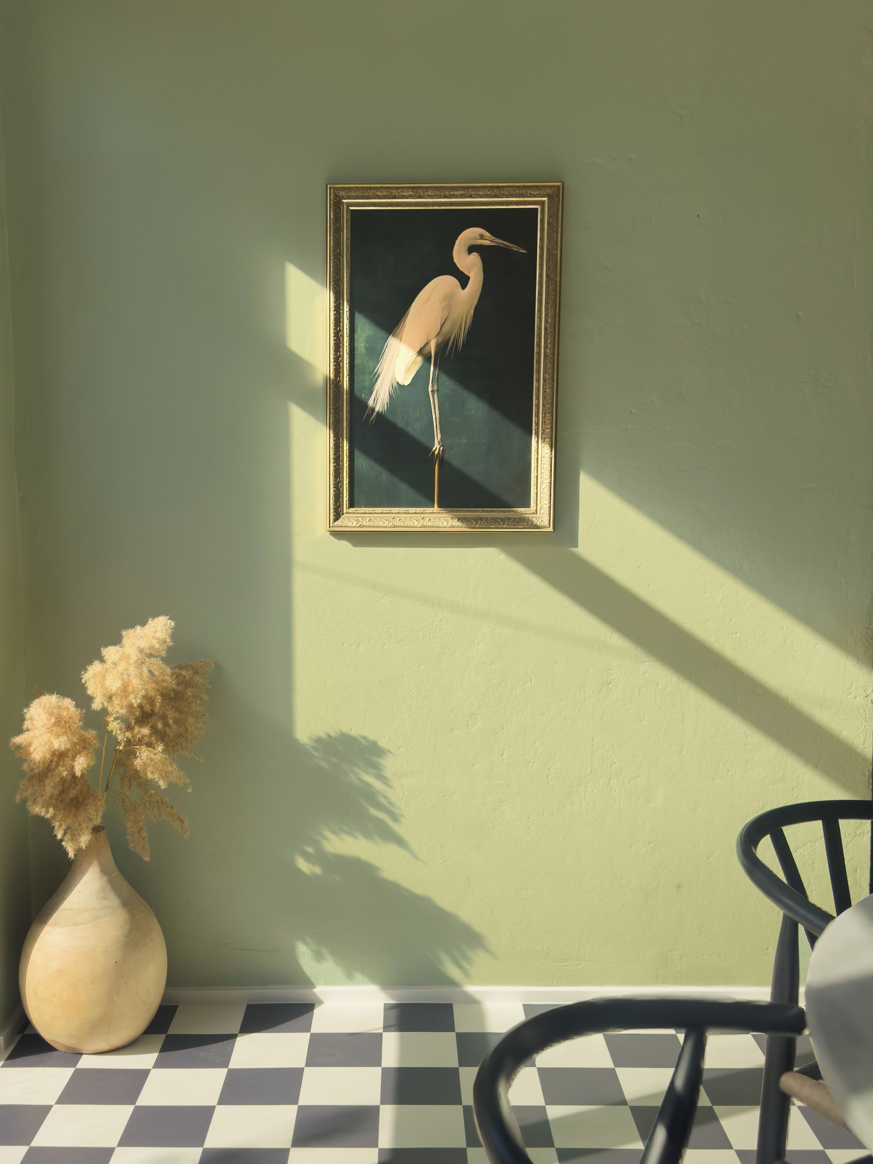 Een lijst met een afbeelding van een vogel aan de muur, een vaas met gedroogde bloemen en een geruite vloer.