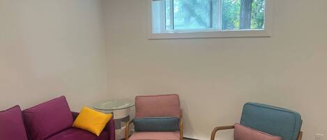 A living room with a purple sofa, a pink armchair, and a blue armchair, a small glass table, and a window with a view of trees.