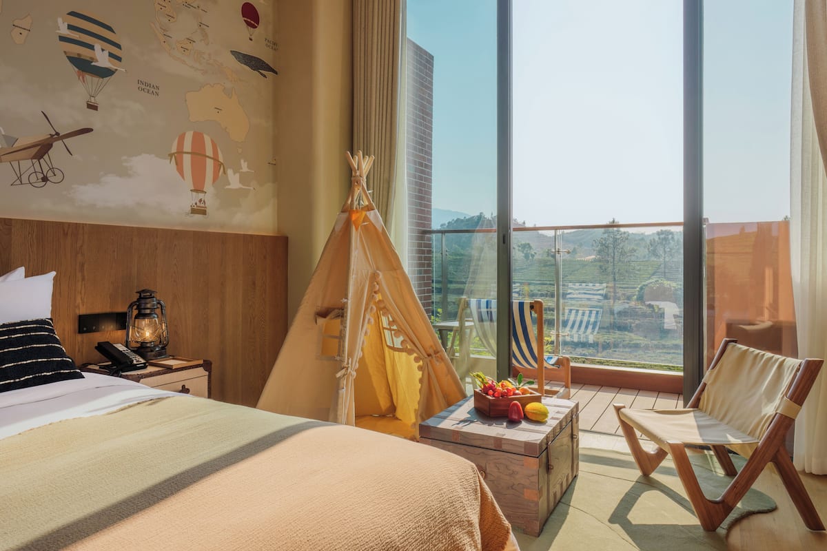Hotelkamer met een bed, een houten nachtkastje, een telefoon, een lantaarn, een tipitent, een fruitmand, een stoel en een groot raam met uitzicht op groen.