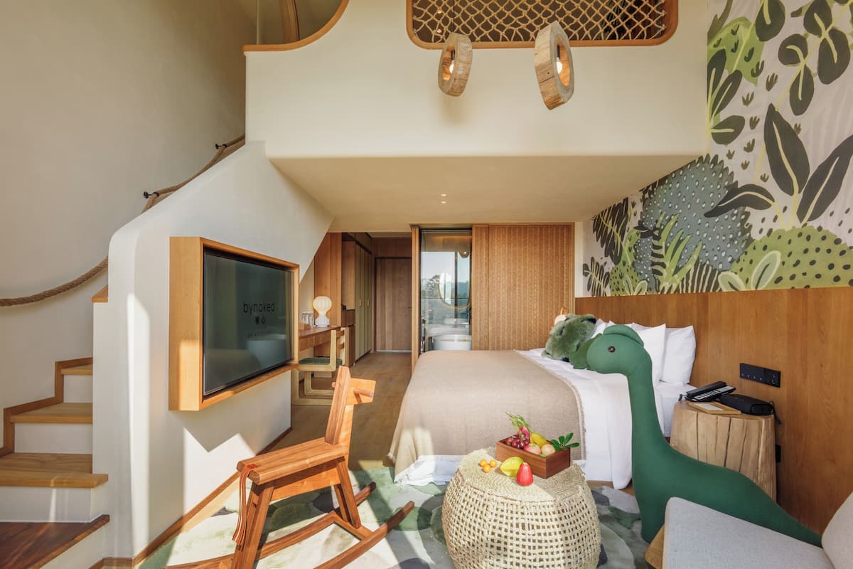 Een moderne hotelkamer met een trap, een bed met een groen dinosaurus kussentje, een televisie en een houten nachtkastje.
