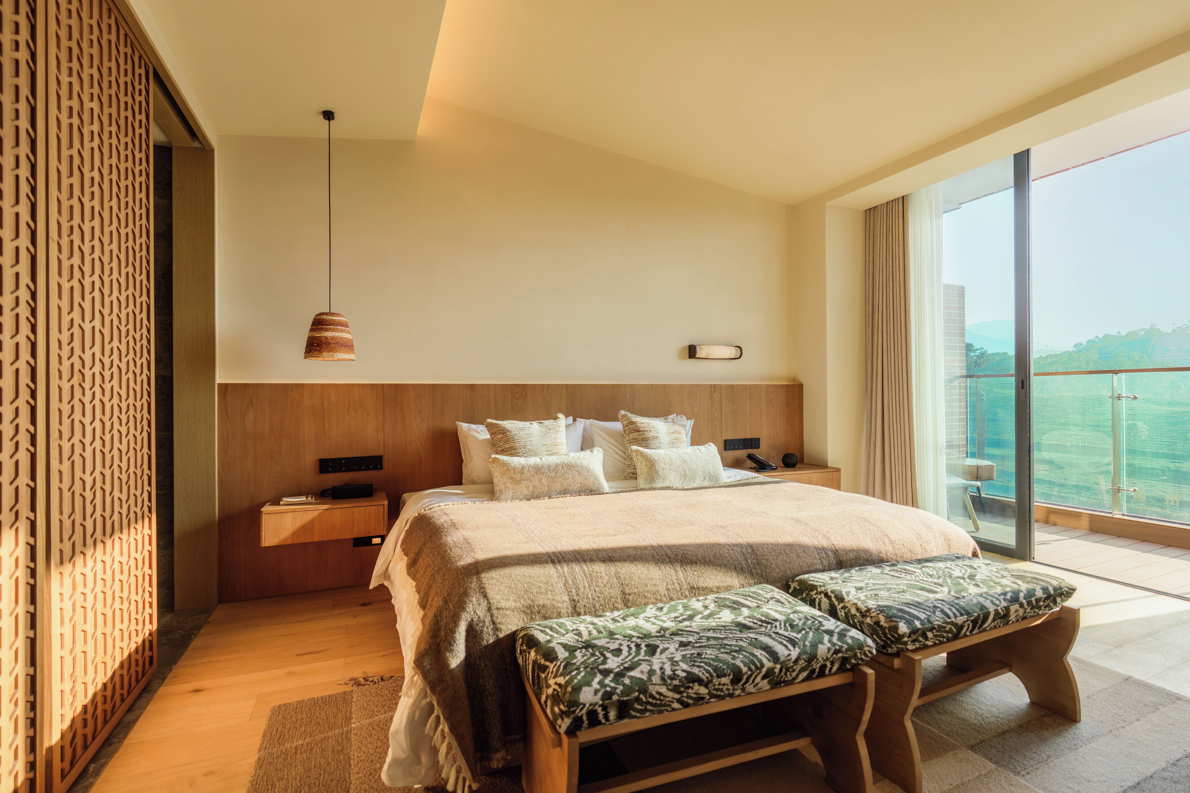A modern bedroom with a large bed, a bedside table, a bench, a hanging light, and a large window with a view.
