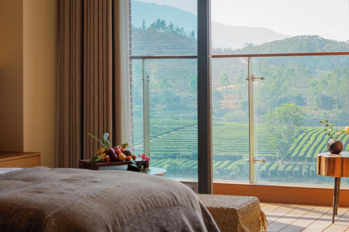 Een slaapkamer met een groot raam, een bed, een nachtkastje met fruit en uitzicht op een groen landschap.