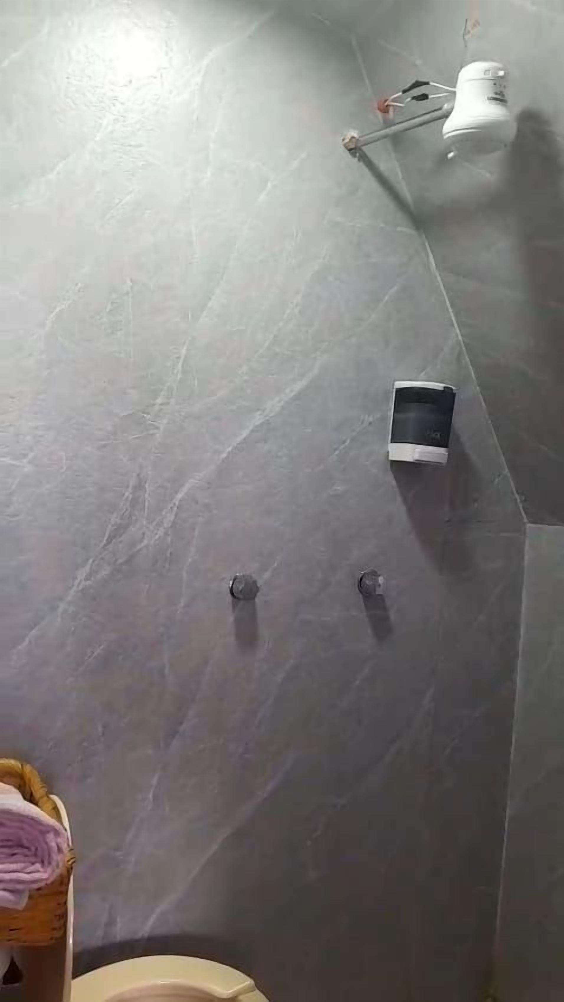 Un baño con piso de baldosas grises, un accesorio de luz blanco montado en la pared, un pequeño dispositivo negro en la pared y una cesta con toallas.