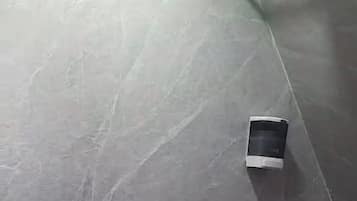Un baño con piso de baldosas grises, un accesorio de luz blanco montado en la pared, un pequeño dispositivo negro en la pared y una cesta con toallas.