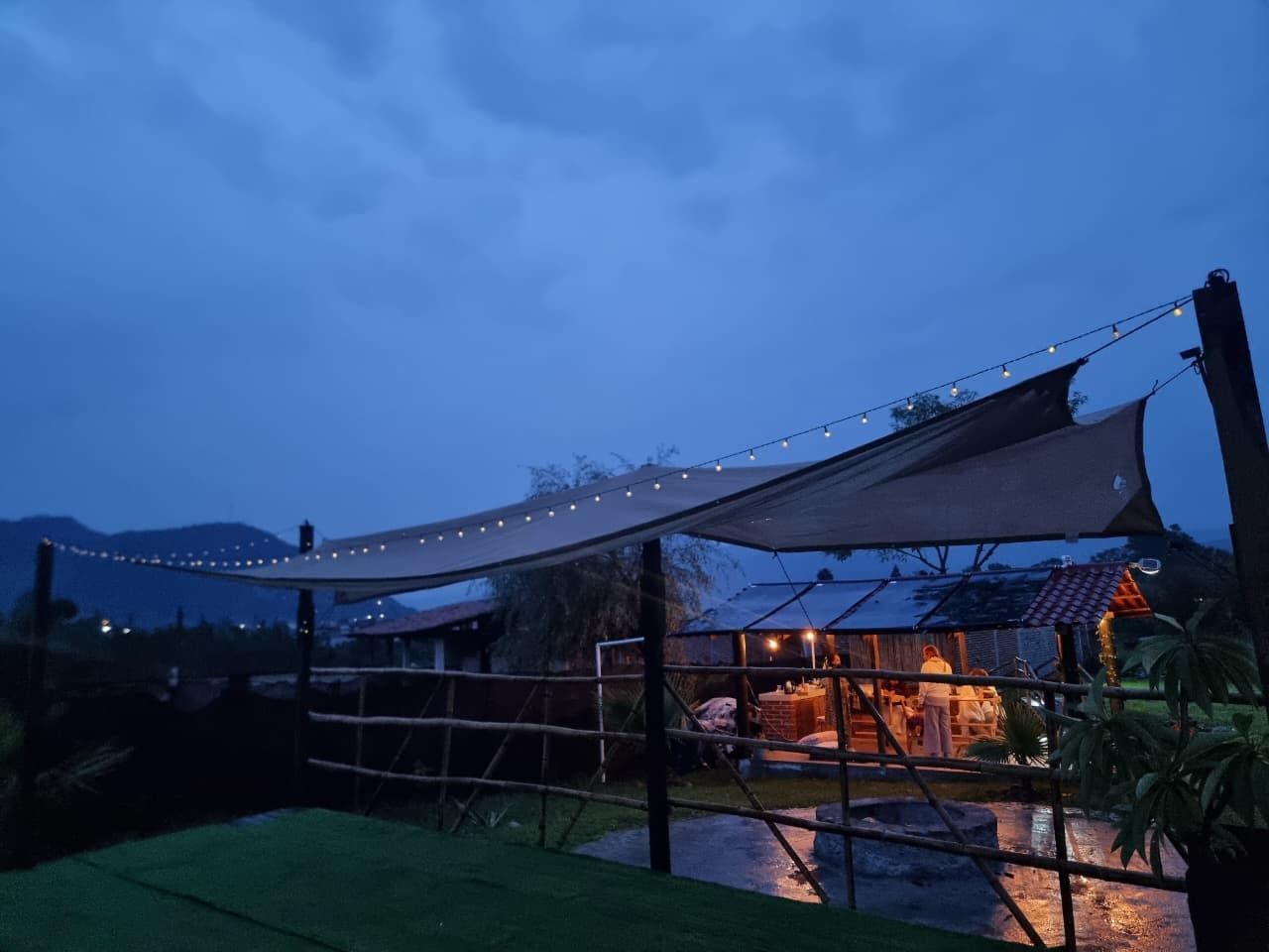 A covered outdoor area with string lights, a shaded seating area, and a building with a tiled roof.