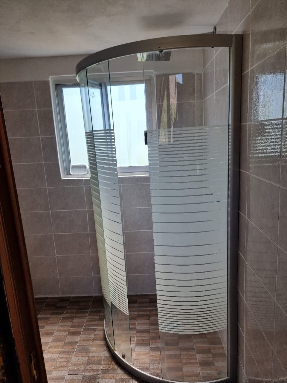 A glass-enclosed shower with a curved design, a window above it, and tiled walls.