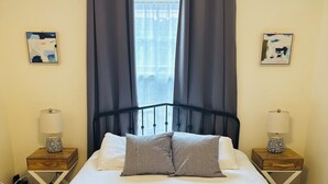 A neatly made bed with pillows, a pair of towels, and a metal headboard. There are two bedside tables with lamps and a window with curtains.
