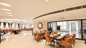 A spacious dining area with round tables and orange chairs, a large mirror on the wall, and a view of the outside through large windows. - White Wharf Manesar (Gurugram)