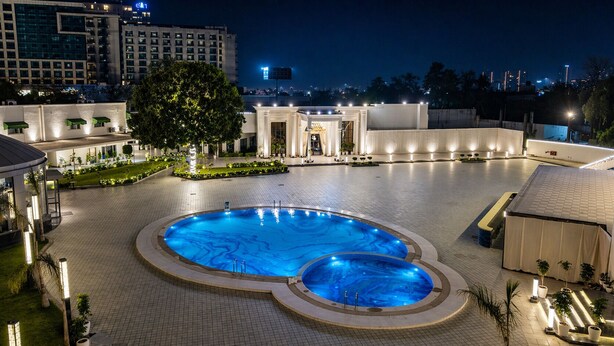 A large indoor swimming pool with a circular design, surrounded by a paved area with potted plants and a building in the background. - White Wharf Manesar (Gurugram)