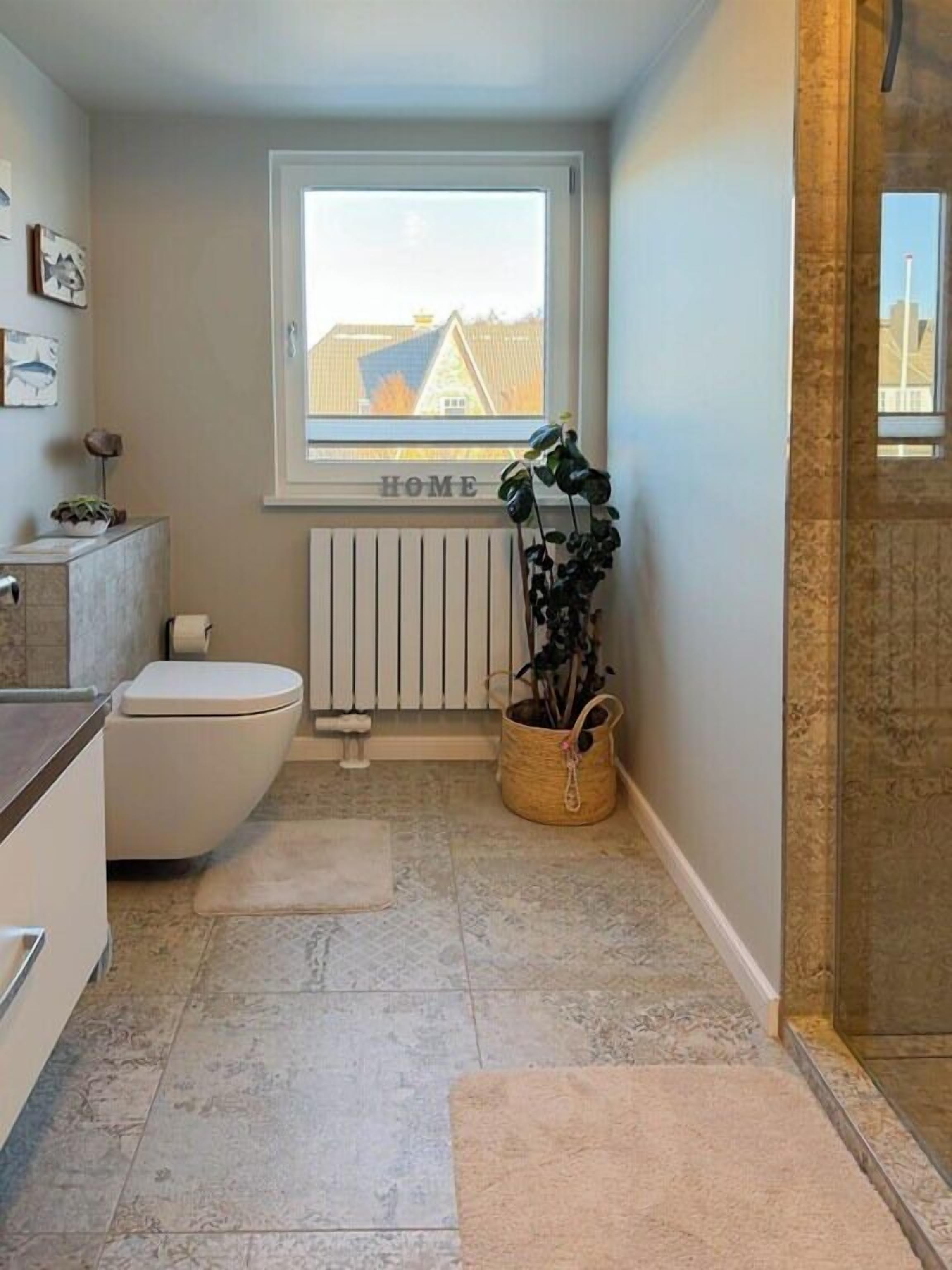 Une petite salle de bain comprenant des toilettes, un radiateur et une fenêtre donnant sur les bâtiments environnants.
