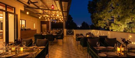 An outdoor dining area with tables set for two, under a covered roof with string lights.
