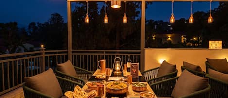 An outdoor dining area with a table set for a meal, surrounded by wicker chairs, under a canopy with string lights.