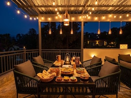 An outdoor dining area with a table set for a meal, surrounded by wicker chairs, under a canopy with string lights.