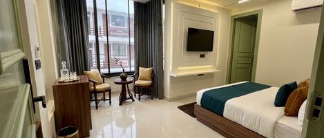 A modern hotel room with a bed, a TV mounted on the wall, a desk with a chair, and a view of the outside.