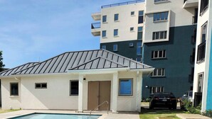 Un edificio residencial con piscina, una estructura pequeña adosada y un edificio de apartamentos de varios pisos al fondo.