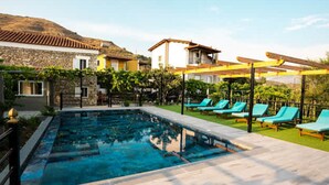 Une piscine entourée de bâtiments en pierre et de verdure, avec des chaises longues et une pergola.
