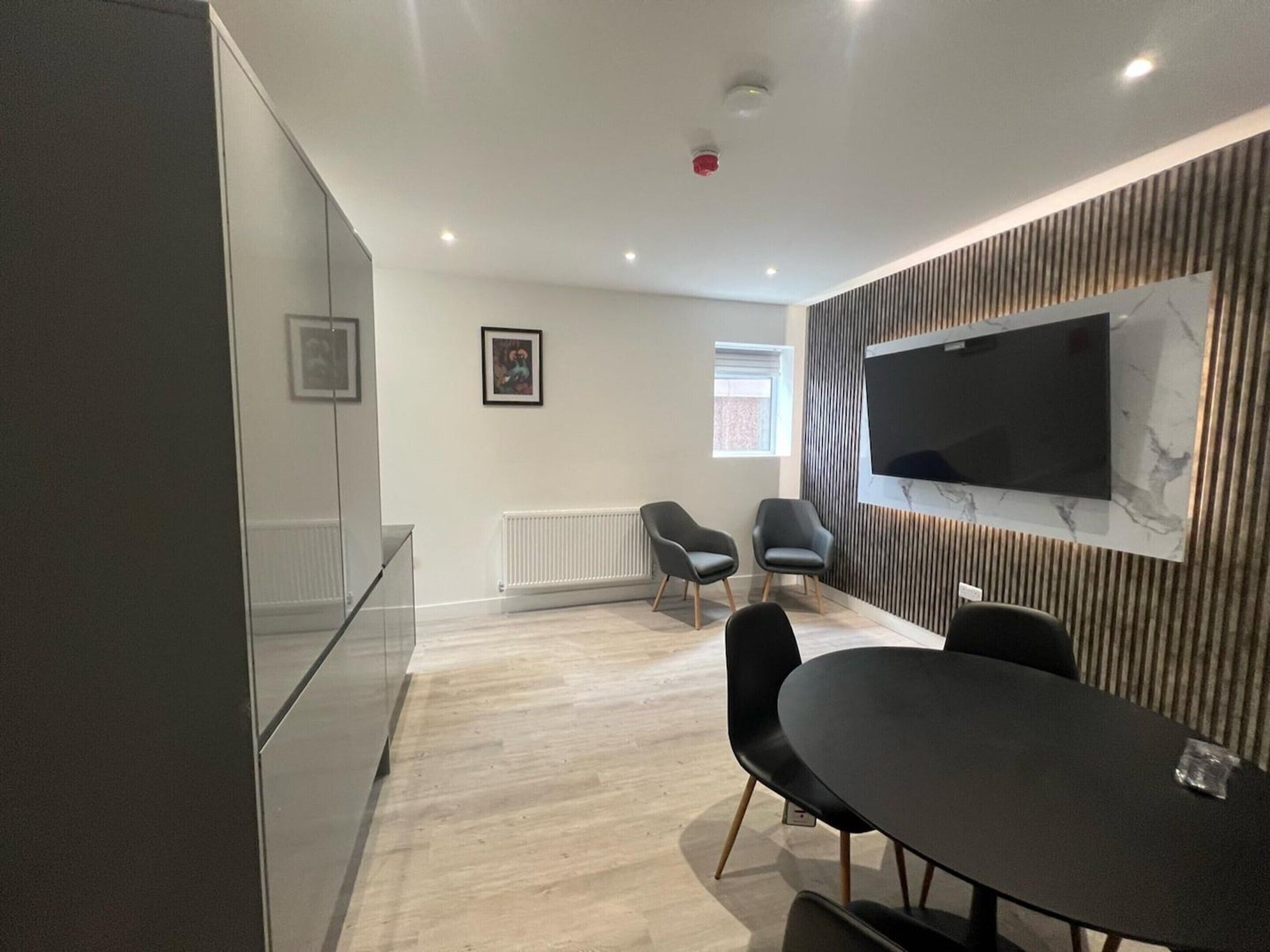 A modern, well-lit room with a large flat-screen TV mounted on a wall, a dining table with chairs, and a radiator on the floor.