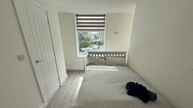 A bedroom with a bed, a window with blinds, a door, and a view of a car outside. - Deluxe Single Room (Manchester)