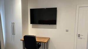 A room with a wooden desk, a black office chair, a flat-screen TV mounted on the wall, a white door, and a wardrobe with silver handles. - Deluxe Single Room (Manchester)