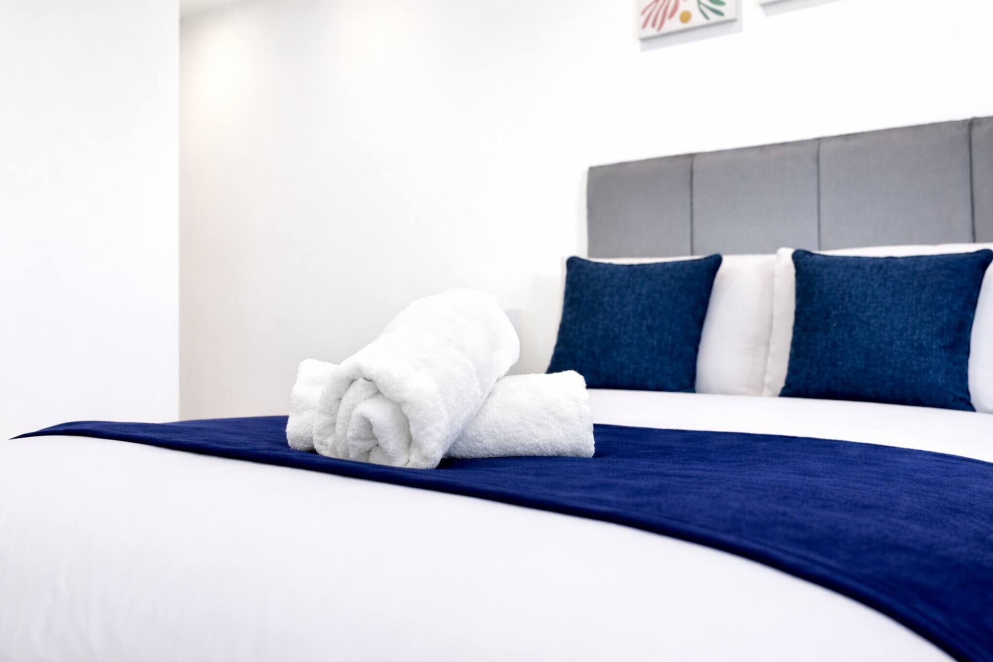 A neatly made bed with a blue bedspread and white pillows, a folded white towel, and a grey headboard.