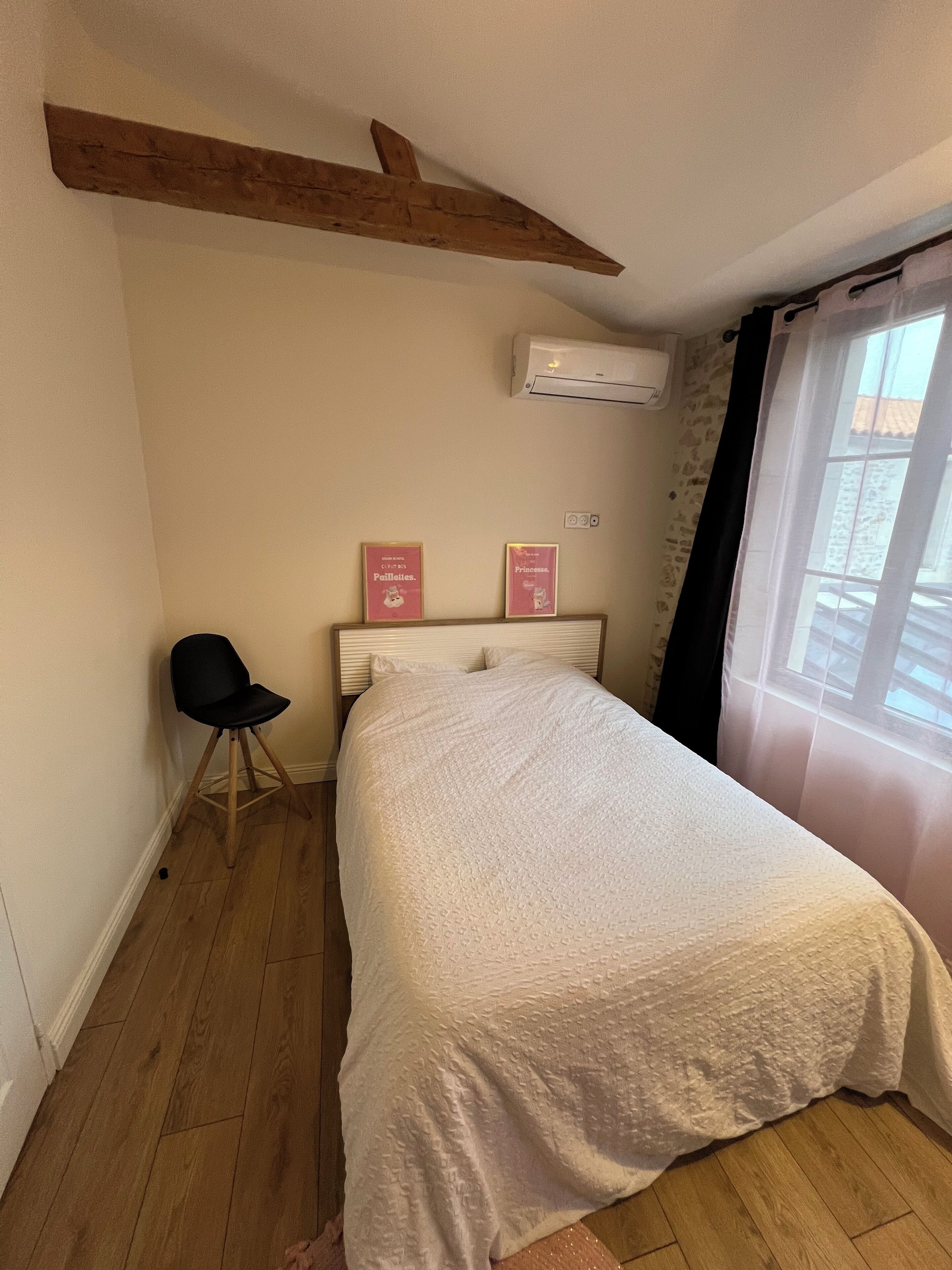 Un dormitorio con cama, una silla, techo con vigas de madera, aire acondicionado y ventana con cortinas.