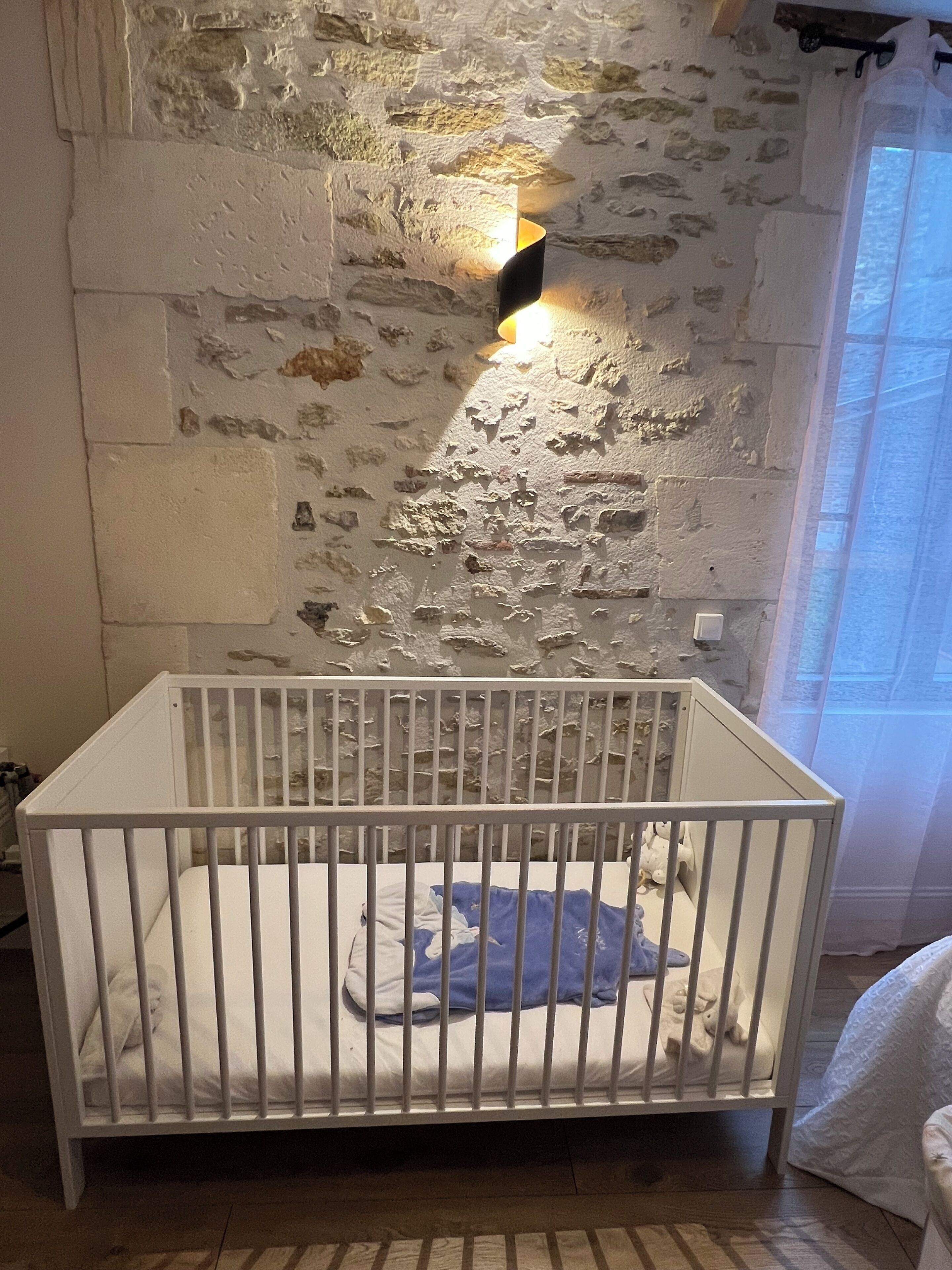 Un berceau blanc est placé contre un mur en pierre à la texture marquée, avec un petit luminaire fixé au-dessus.