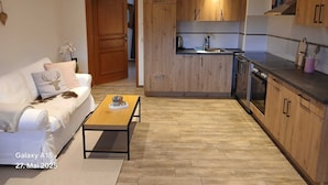 Una cucina moderna con mobili in legno, forno a incasso e lavello. Nell'area soggiorno, un divano bianco con un cuscino decorativo e un piccolo tavolo in legno.