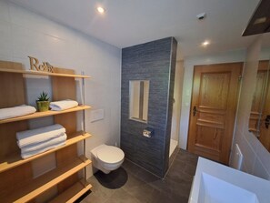 Ein modernes Badezimmer mit Toilette, Regalen mit gefalteten Handtüchern, einer kleinen Topfpflanze und einer Holztür.